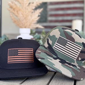 Puede incluir: Dos gorras de camionero con parches de la bandera estadounidense. Una gorra es negra con una bandera de color cobre, y la otra es de camuflaje con una bandera blanca y negra. Ambas tienen la parte trasera de malla.