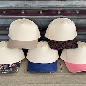 以下が含まれることがあります： クリーム色のクラウンと様々な色のつばが付いた6つの野球帽のコレクション。つばには、ピンクとブルーなどの単色、ヒョウ柄、迷彩柄、無地のタンカラーが含まれています。
