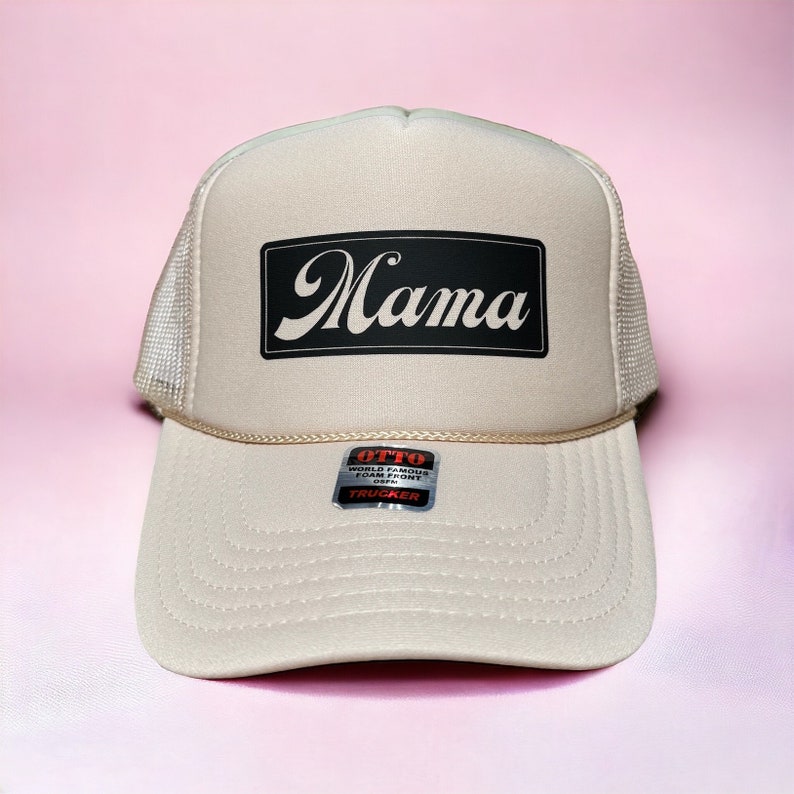 May include: A beige trucker hat with a black patch that says "Mama" in a stylized font. The hat has a mesh back and a white adjustable strap.