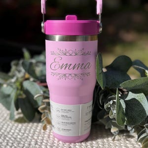 May include: A pink insulated tumbler with a matching handle and lid. The tumbler features the name "Emma" in a decorative font, surrounded by floral accents. The tumbler is designed to hold 32 ounces.