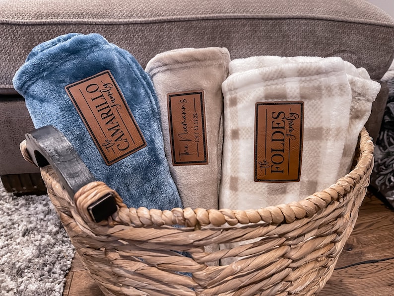 May include: Three blankets in a wicker basket. The blankets are blue, beige, and white with a plaid pattern. Each blanket has a brown leather label with the words 'The Camarillo Family', 'The McHenry's Est. 11.11.22', and 'The Foldes Family'.