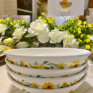 Könnte beinhalten: Ein Satz von vier weißen Keramikschalen mit einem Sonnenblumen- und Grünblattdesign. Die Schalen sind übereinander gestapelt. Die Schalen befinden sich auf einer weißen Oberfläche. Eine Karte mit dem Text "MULUYA ESSENTIALS LLC" befindet sich im Hintergrund.