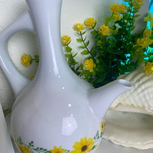 Puede incluir: Jarra de cerámica blanca con asa y boquilla. La jarra está decorada con girasoles amarillos y hojas verdes. Tiene una forma y un diseño únicos, adecuada para servir bebidas.