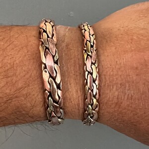May include: Two braided bracelets made of copper, silver, and gold metal. The bracelets are worn on a person's wrist.