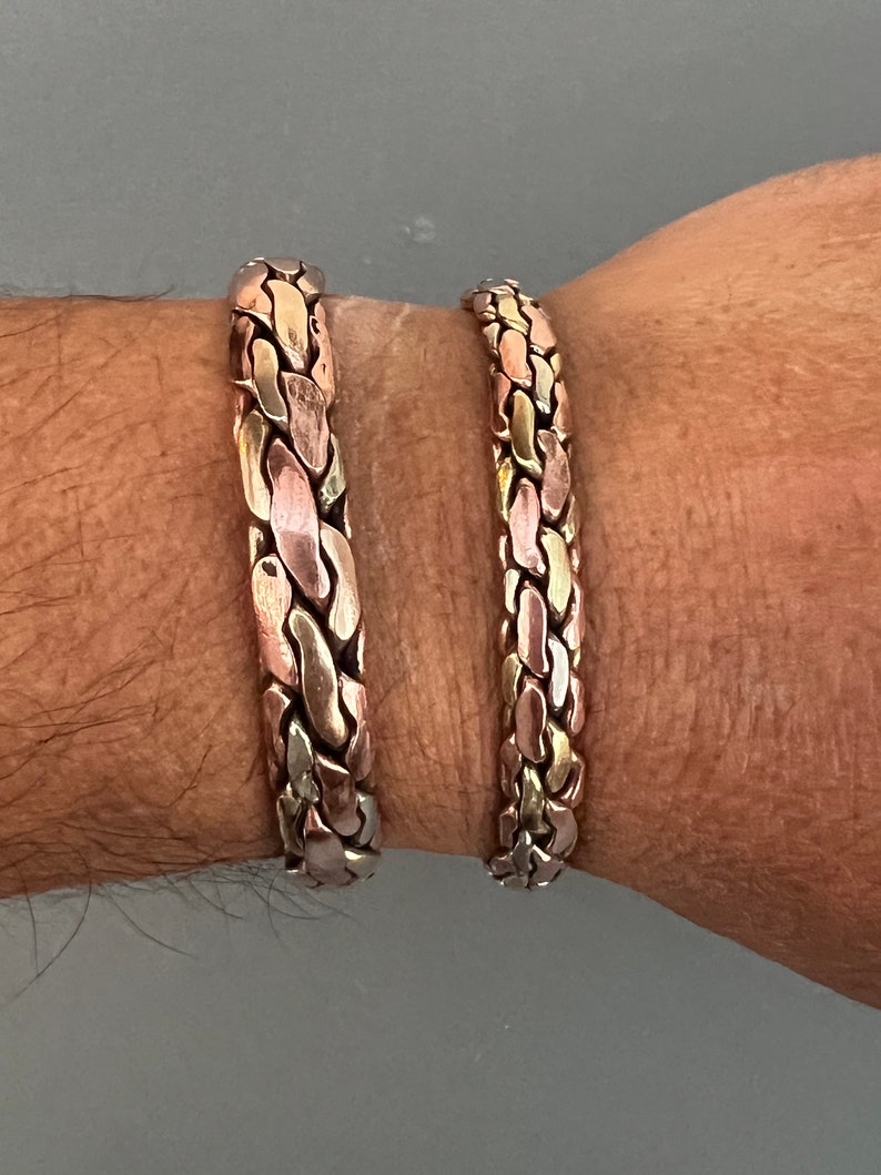 May include: Two braided bracelets made of copper, silver, and gold metal. The bracelets are worn on a person's wrist.