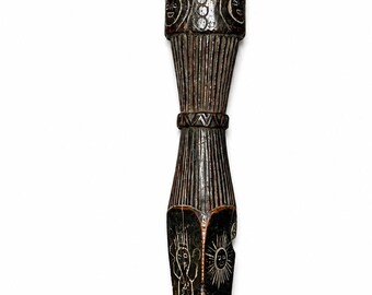 May include: A dark brown carved wooden object with a tapered, pointed shape. The surface features intricate white etched designs including suns, figures, and geometric patterns. A frayed red fabric strip is attached to the top with metal pins.