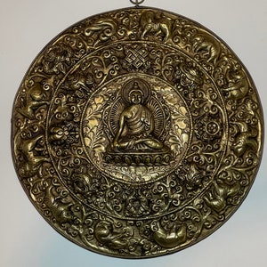 May include: A round, gold-colored metal wall hanging with a raised image of a seated Buddha surrounded by intricate floral and animal designs.