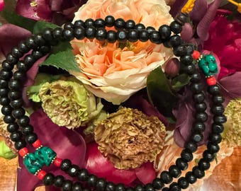 Puede incluir: Un collar largo de cuentas de madera de color marrón oscuro dispuestas en un bucle circular. La pieza presenta dos pequeños acentos de piedra verde tallada y cuentas espaciadoras de color rojo brillante, con un nudo de cordón amarillo.