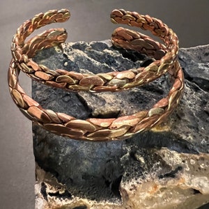 May include: Two braided copper bracelets with a hammered finish. The bracelets are stacked on top of each other and are resting on a rock.
