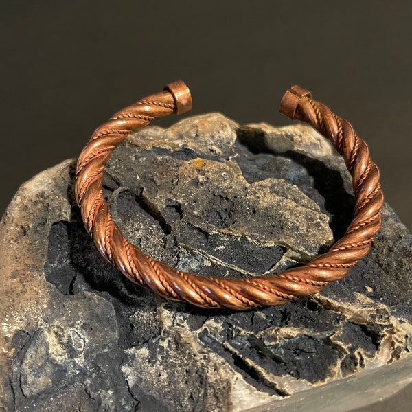 May include: A copper bracelet with a twisted design. The bracelet is on a rough, gray rock.