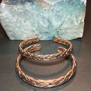 May include: Two braided copper bracelets with a silver and gold finish. The bracelets are stacked on top of each other.