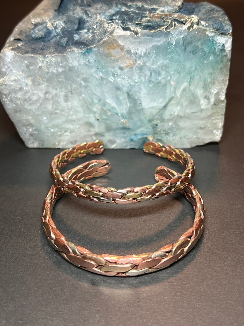 May include: Two braided copper bracelets with a silver and gold finish. The bracelets are stacked on top of each other.