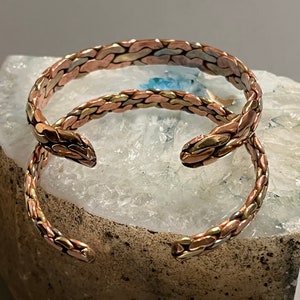 May include: Two braided copper bracelets with a mix of gold and silver tones. The bracelets are stacked on top of each other and are resting on a white and gray textured surface.