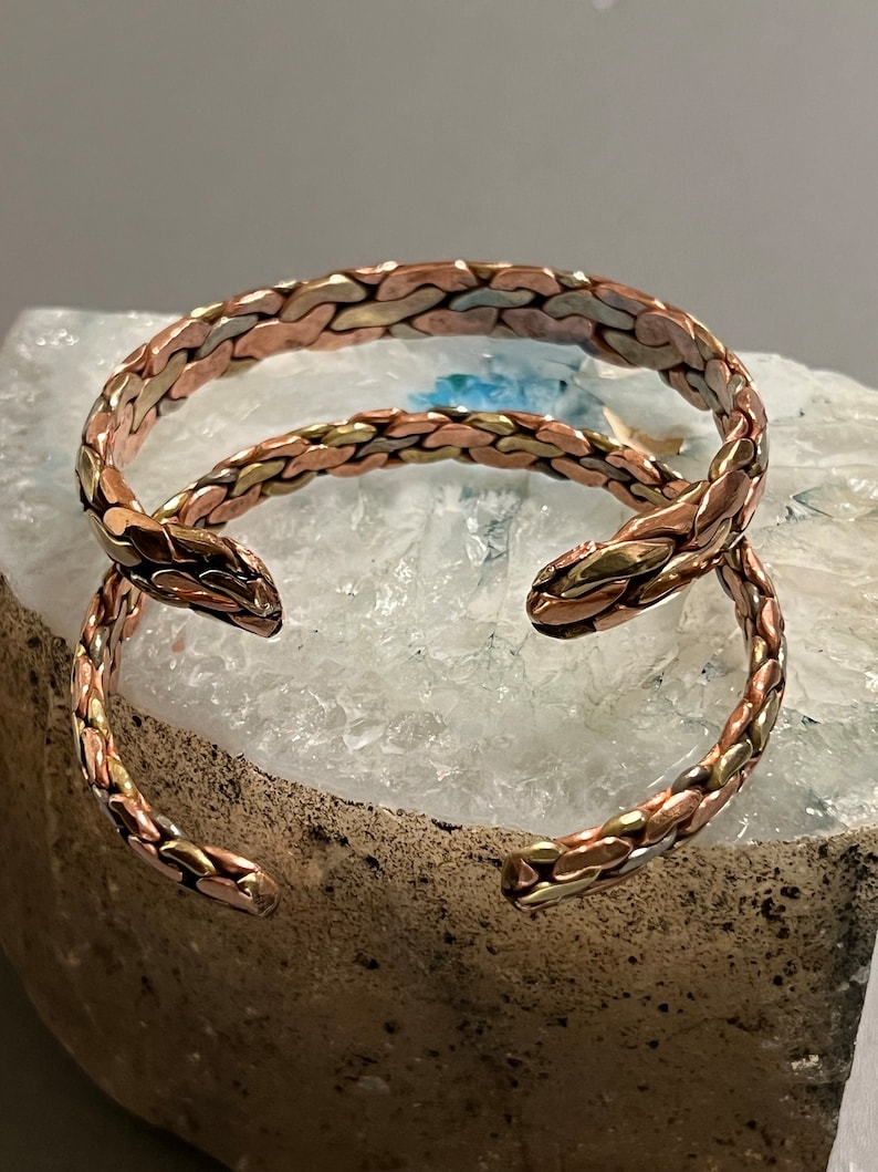 May include: Two braided copper bracelets with a mix of gold and silver tones. The bracelets are stacked on top of each other and are resting on a white and gray textured surface.