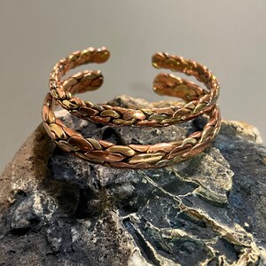 May include: Two braided copper bracelets with a mixed metal finish. The bracelets are stacked on top of each other and are resting on a gray rock.