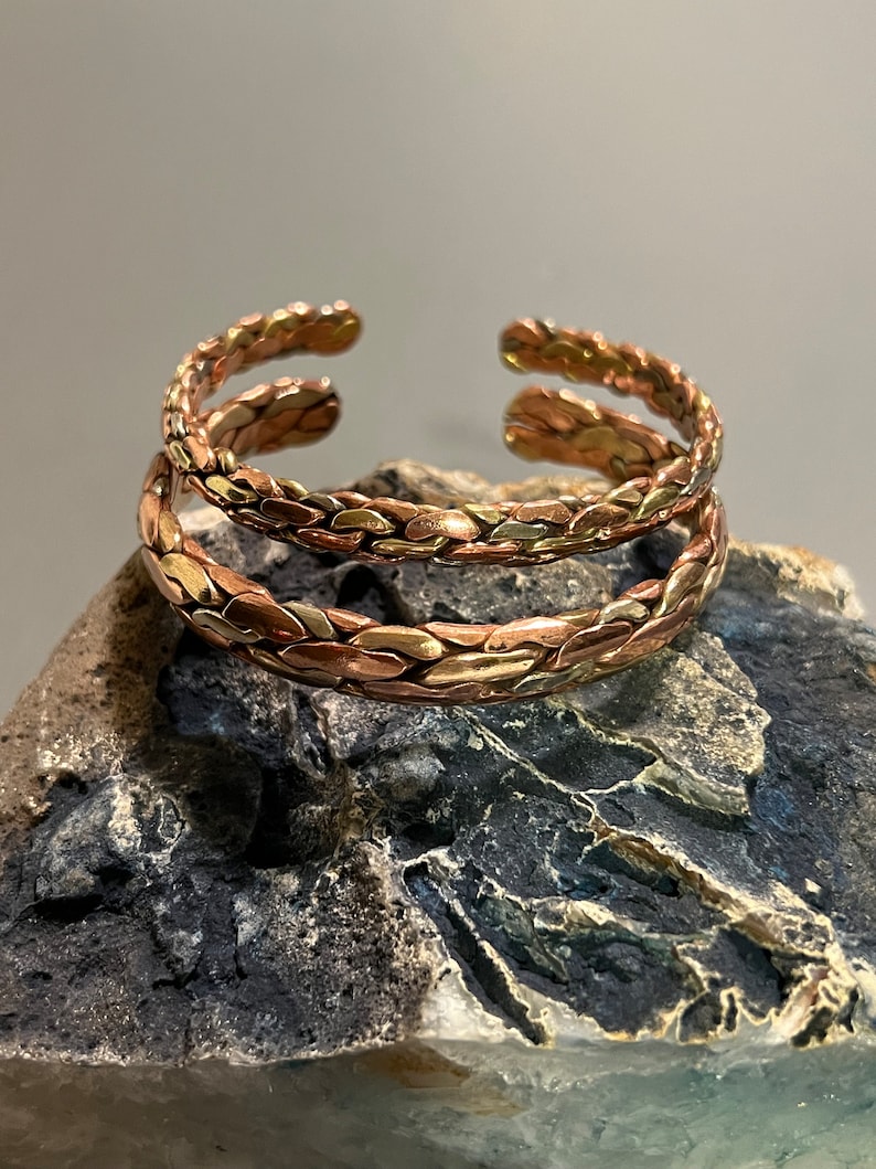 May include: Two braided copper bracelets with a mixed metal finish. The bracelets are stacked on top of each other and are resting on a gray rock.