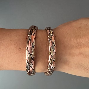 May include: Two braided copper bracelets with a mix of silver and copper tones. The bracelets are worn on a person's wrist.