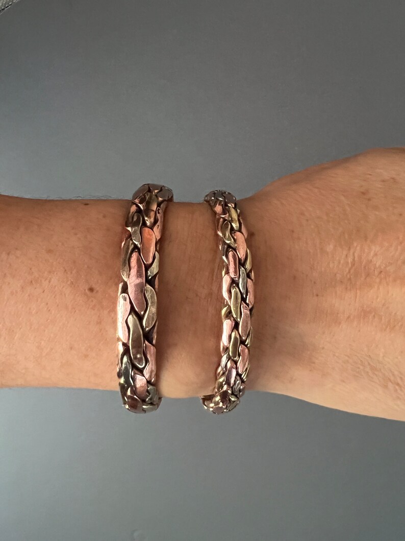 May include: Two braided copper bracelets with a mix of silver and copper tones. The bracelets are worn on a person's wrist.