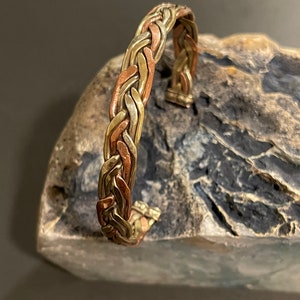 May include: A braided bracelet made of three different metals: gold, copper, and silver. The bracelet is open and adjustable to fit different wrist sizes.