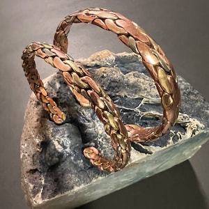 May include: Two braided copper bracelets with a hammered finish. The bracelets are stacked on top of each other and have a warm, metallic sheen.