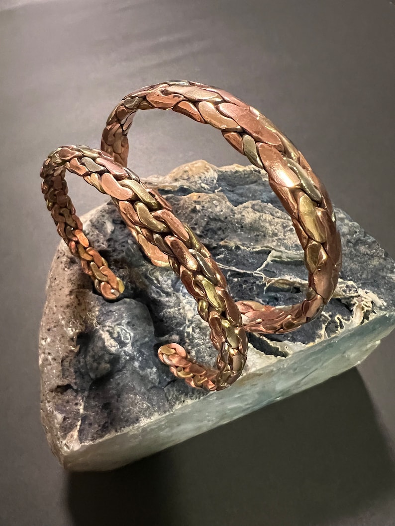 May include: Two braided copper bracelets with a hammered finish. The bracelets are stacked on top of each other and have a warm, metallic sheen.