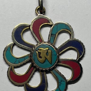 May include: A silver pendant in the shape of a flower with a turquoise, red, and blue inlay. The center of the flower has a gold Om symbol on a turquoise background.