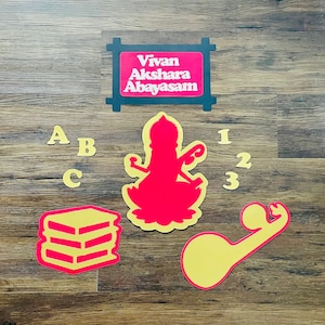 May include: A collection of colorful cutouts on a wood-grain surface. Includes the text "Vivan Akshara Abayasam" in a framed sign, letters A, B, and C, numbers 1, 2, and 3, a stack of books, and a figure in a lotus position.