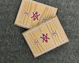 May include: Two wooden coasters with a red star and white geometric designs on a brown background. The coasters have a white border with red dots.