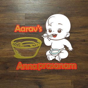 May include: A gold-colored bowl with a white baby illustration holding a spoon and a bowl. The text "Aarav's Annapraasanam" is in red.