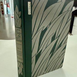 May include: A hardcover book titled The Jungle Books Vol 2 by Kipling, featuring a dark green spine with gold leaf illustrations and text, and a cover with a grey leaf pattern. A white rectangular label with the word JOURNAL is on the front.