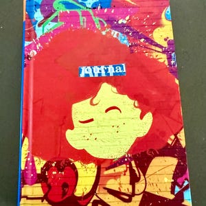 May include: A rectangular hardcover journal featuring a vibrant graffiti-style cover with a brick wall pattern in shades of red, yellow, and blue. The center displays a stylized illustration of a character with red hair and the word Journal.