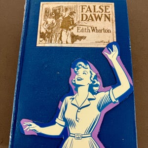May include: A blue hardcover book titled False Dawn by Edith Wharton with a rectangular illustration on the cover. A die-cut paper cutout of a person in a collared dress with arms raised is layered over the lower portion of the book cover.