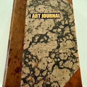 May include: A rectangular book titled ART JOURNAL with a marbled beige and black cover pattern. The spine and corners are bound in a smooth, mottled brown material. The text ART JOURNAL is printed in bold black letters on a yellow rectangular label.