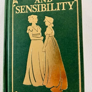May include: A green hardcover journal titled Sense and Sensibility by Jane Austen. The cover features gold foil lettering and a gold silhouette illustration of two figures in long dresses. A small white label with the word journal is attached.