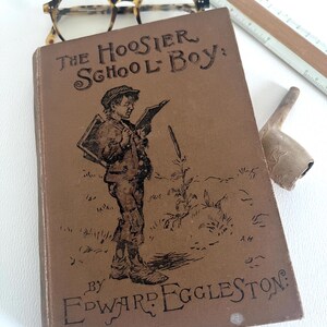 May include: The cover of a vintage book titled "The Hoosier School-Boy" by Edward Eggleston. The cover features a black and white illustration of a boy reading a book.