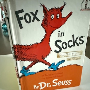 May include: A copy of "Fox in Socks" by Dr. Seuss. The book cover shows a red fox character wearing blue socks. The title is in large black letters. The book is part of the Beginner Books series.