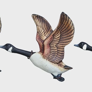 May include: Three decorative goose figurines in mid-flight. Each goose has a black head and neck, white body, and brown and tan wings. The wings are spread wide, and the geese appear to be in motion, with their feet tucked up.