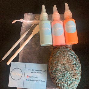 May include: A set of three small bottles of paint in light blue, pink, and orange, with the words "Pick & Pop" on each bottle. The bottles are in a white mesh bag with a small white tool and a blue and brown sponge. The bag also includes a blue card with the words "Pick & Pop" and the text "Designed to clear your thoughts and calm the mind" along with social media handles.