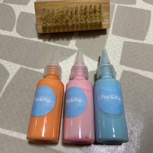 May include: Three small bottles of paint in light blue, pink, and orange. Each bottle has a white label with the text "Pick & Pop" in blue.