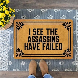 Könnte beinhalten: Eine braune Fußmatte mit schwarzem Text, der "I see the assassins have failed" lautet. Die Fußmatte hat einen dekorativen Rand mit einem floralen Muster.