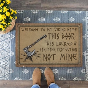 Puede incluir: Una alfombra de bienvenida marrón con un gráfico de hacha vikinga y el texto "Bienvenido a mi hogar vikingo. Esta puerta está cerrada para tu protección, no para la mía."