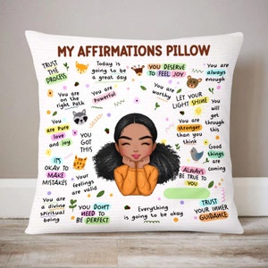 Könnte beinhalten: Ein weißes Kissen mit einem farbenfrohen Design, das ein Cartoon-Mädchen mit Afro-Haaren und positive Affirmationen zeigt. Der Text enthält Sätze wie "Vertraue dem Prozess", "Du bist mächtig", "Du verdienst es, Freude zu empfinden", "Lass dein Licht scheinen", "Du hast das", "Du musst nicht perfekt sein", "Sei immer du selbst" und "Vertraue deiner inneren Führung".