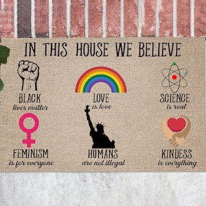 Puede incluir: Una alfombra de bienvenida con el texto "IN THIS HOUSE WE BELIEVE" e imágenes que representan varios temas de justicia social e igualdad. Las imágenes incluyen un puño en alto, un arcoíris, un símbolo científico, un símbolo femenino, la Estatua de la Libertad y un corazón con manos alrededor. El texto debajo de cada imagen dice "BLACK lives matter", "LOVE is love", "SCIENCE is real", "FEMINISM is for everyone", "HUMANS are not illegal", y "KINDNESS is everything".