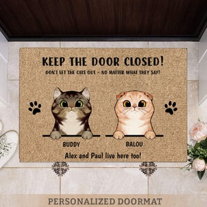 May include: A brown doormat with the text "KEEP THE DOOR CLOSED! DON'T LET THE CATS OUT - NO MATTER WHAT THEY SAY!" and two cartoon cats with the names "BUDDY" and "BALOU" above them. The text "Alex and Paul live here too!" is below the cats. The doormat has a brown burlap texture.
