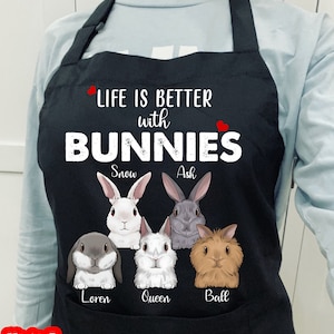 Peut inclure: Tablier noir avec le texte "Life is better with bunnies" et cinq lapins avec les noms "Snow", "Ash", "Loren", "Queen" et "Ball".