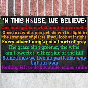 May include: Black sign with white text that reads "IN THIS HOUSE, WE BELIEVE:" followed by a list of sayings. The sign has a rainbow border.