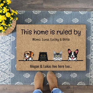 Könnte beinhalten: Eine braune Fußmatte mit dem Text "This home is ruled by Momo, Luna, Lucky & Bella. Megan & Lukas live here too..." und Illustrationen eines Beagles, zweier Katzen und eines Corgis.