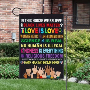 Puede incluir: Una bandera de jard&iacute;n negra con el texto "En esta casa creemos: Black Lives Matter, Love is Love, Los derechos de las mujeres son derechos humanos, La ciencia es real, Ning&uacute;n humano es ilegal, La amabilidad lo es todo, En la libertad religiosa, El odio no tiene hogar aqu&iacute;." La bandera tambi&eacute;n presenta un coraz&oacute;n arco&iacute;ris, un signo de paz arco&iacute;ris, un pu&ntilde;o arco&iacute;ris, una mano arco&iacute;ris y un coraz&oacute;n arco&iacute;ris con un signo de paz. La bandera est&aacute; colgada en un poste de metal.