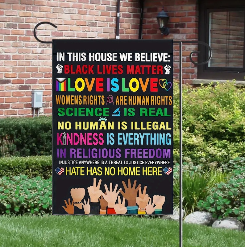 Puede incluir: Una bandera de jard&iacute;n negra con el texto "En esta casa creemos: Black Lives Matter, Love is Love, Los derechos de las mujeres son derechos humanos, La ciencia es real, Ning&uacute;n humano es ilegal, La amabilidad lo es todo, En la libertad religiosa, El odio no tiene hogar aqu&iacute;." La bandera tambi&eacute;n presenta un coraz&oacute;n arco&iacute;ris, un signo de paz arco&iacute;ris, un pu&ntilde;o arco&iacute;ris, una mano arco&iacute;ris y un coraz&oacute;n arco&iacute;ris con un signo de paz. La bandera est&aacute; colgada en un poste de metal.