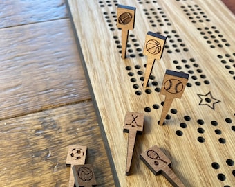 Può includere: Tavolo da gioco in legno con una stella e dei buchi. Il tavolo ha dei pioli in legno con diverse icone sportive, tra cui una palla da golf, un pallone da calcio, una pallavolo, una pallacanestro, una palla da baseball, bastoni da hockey e una racchetta da tennis.